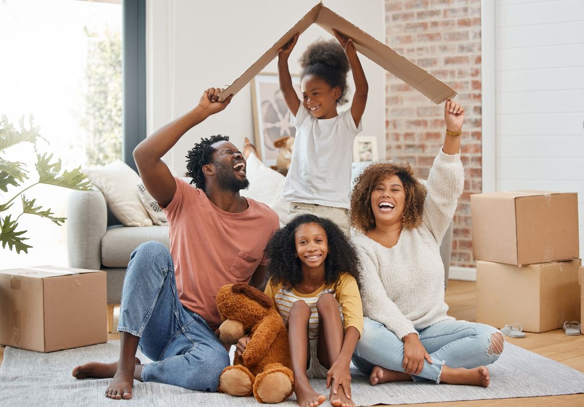 A family is sitting on the floor in a living room holding a cardboard house.