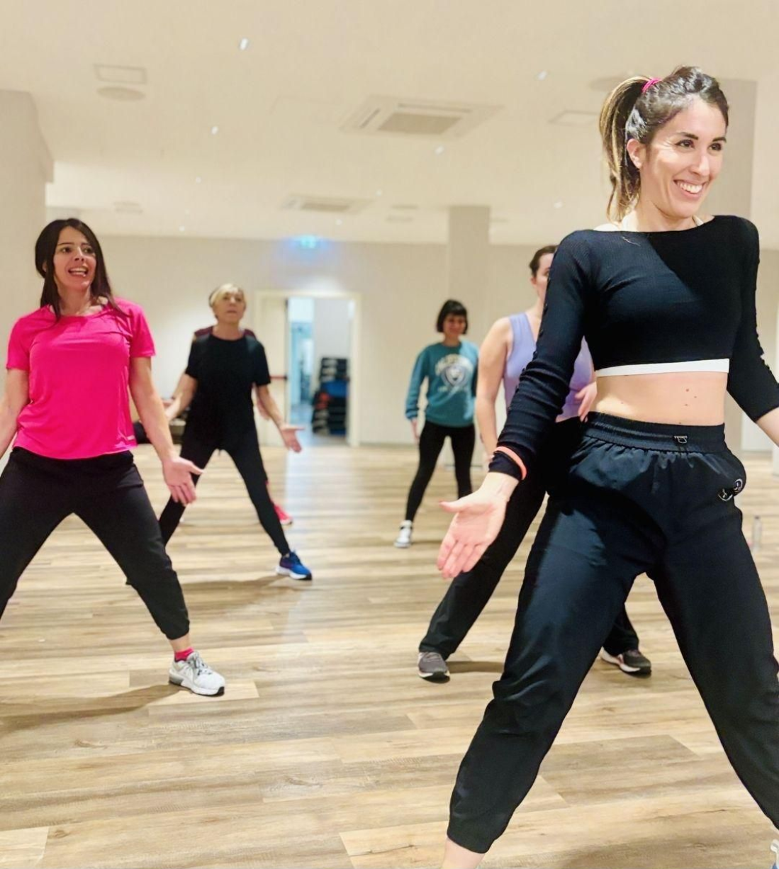 Un gruppo di persone che seguono una lezione di danza in uno studio luminoso; sorridono e indossano abiti da ginnastica.
