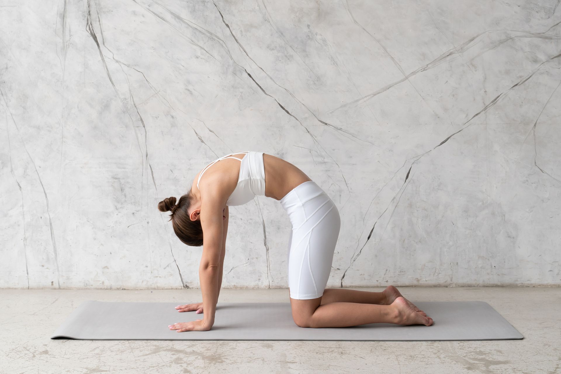 Donna in tenuta da yoga bianca nella posizione del gatto su un tappetino grigio.