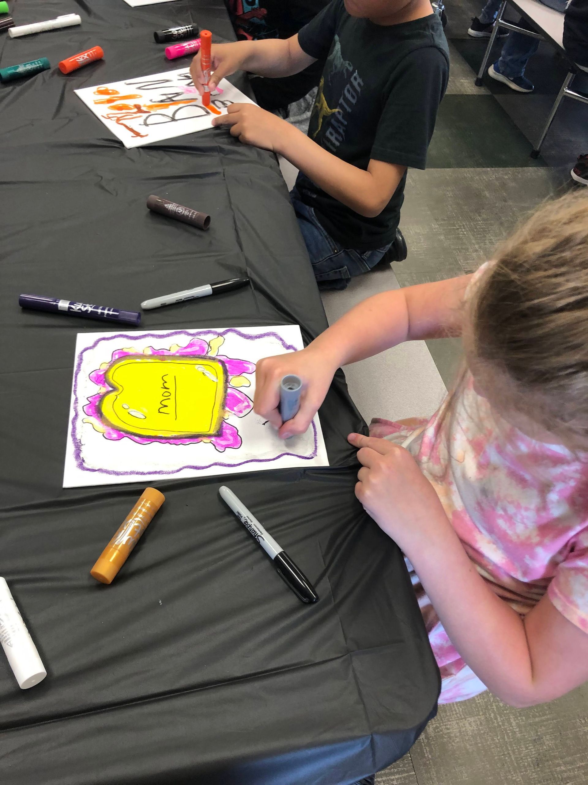 Two children coloring with markers at a table covered in a black cloth.