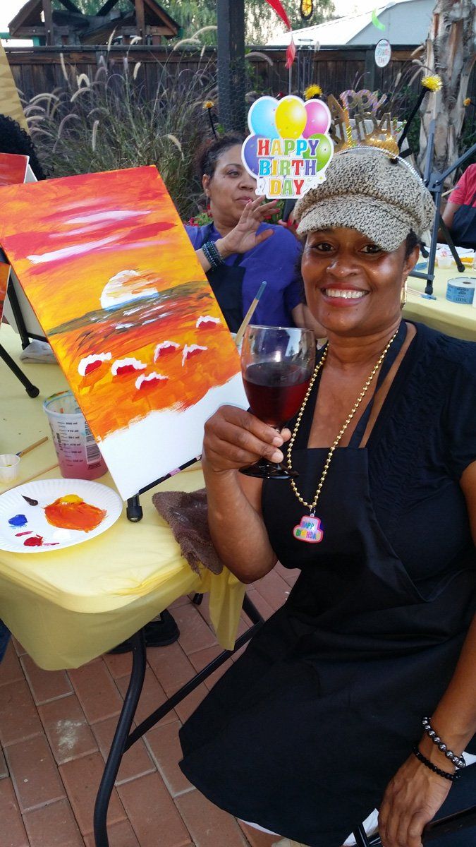 Woman holding wine, smiling, next to a painted sunset canvas during a party; colorful balloons in the background.