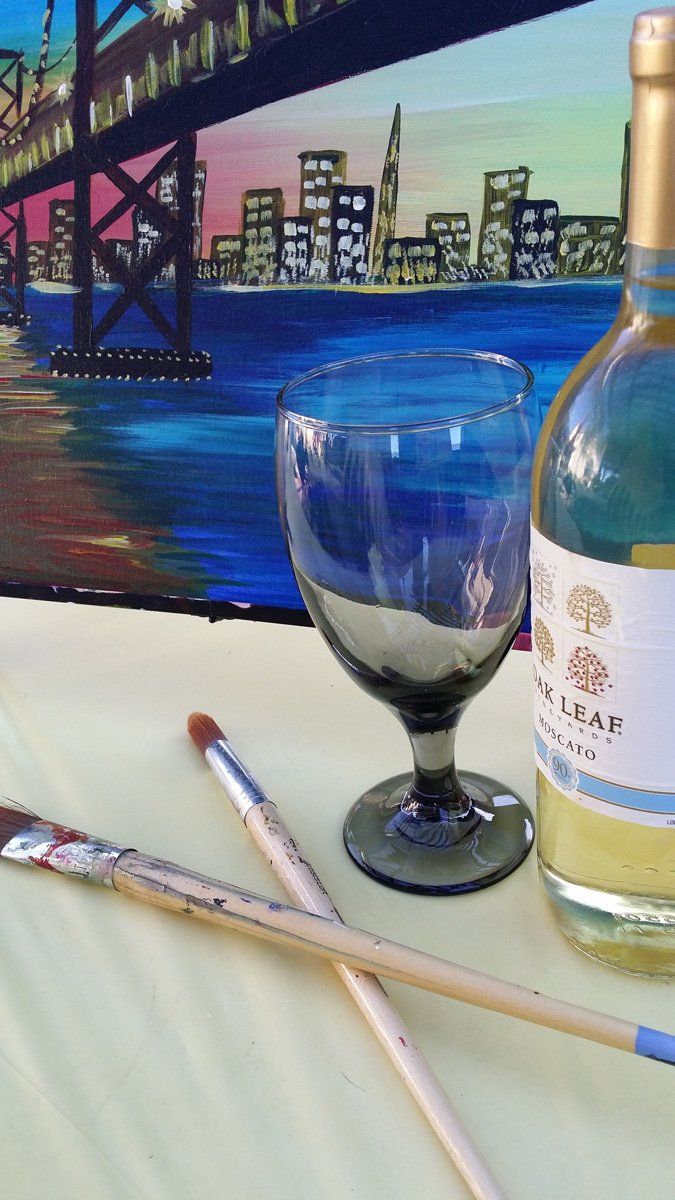 Paintbrushes, a wine glass, and a bottle of wine in front of a painting of a bridge and city skyline.