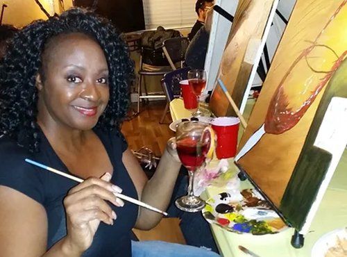 Woman with curly hair painting at easel, holding paintbrush, with glass of wine, smiling.