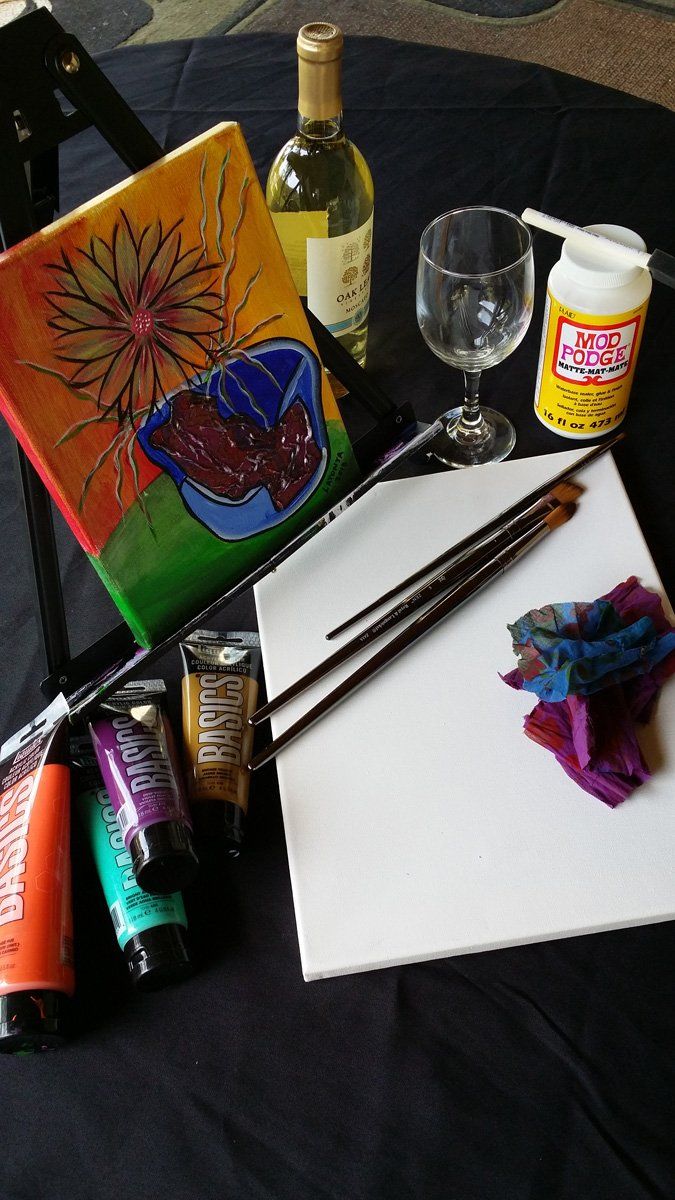Painting supplies arranged on a black tablecloth: canvas, paints, brushes, wine, and a glass.