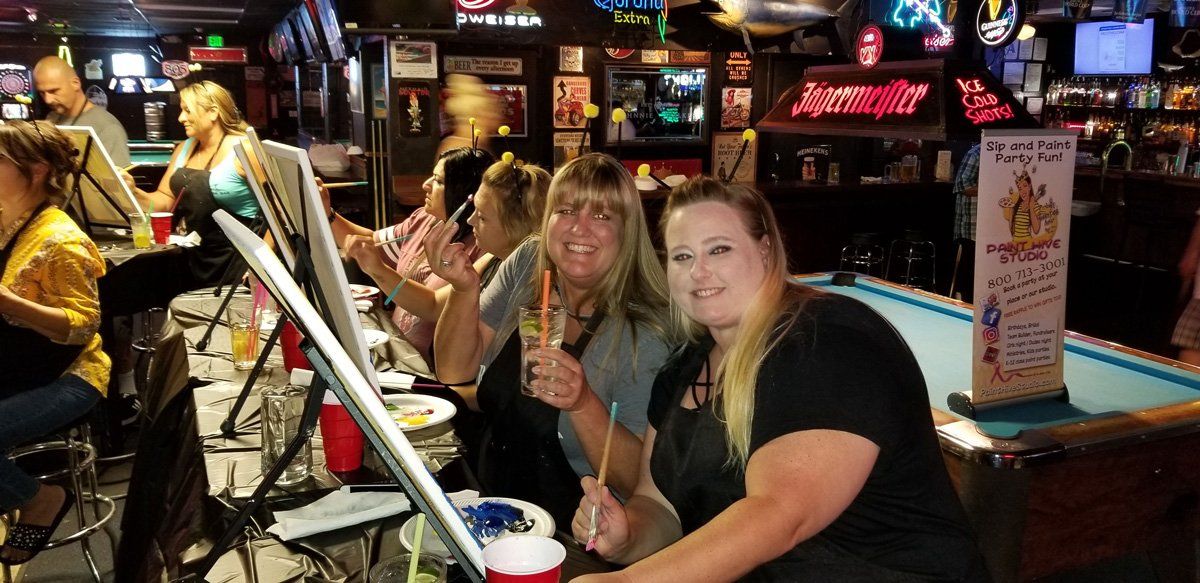 People paint at a bar, some hold drinks, smiling. The setting is dimly lit with pool table visible.