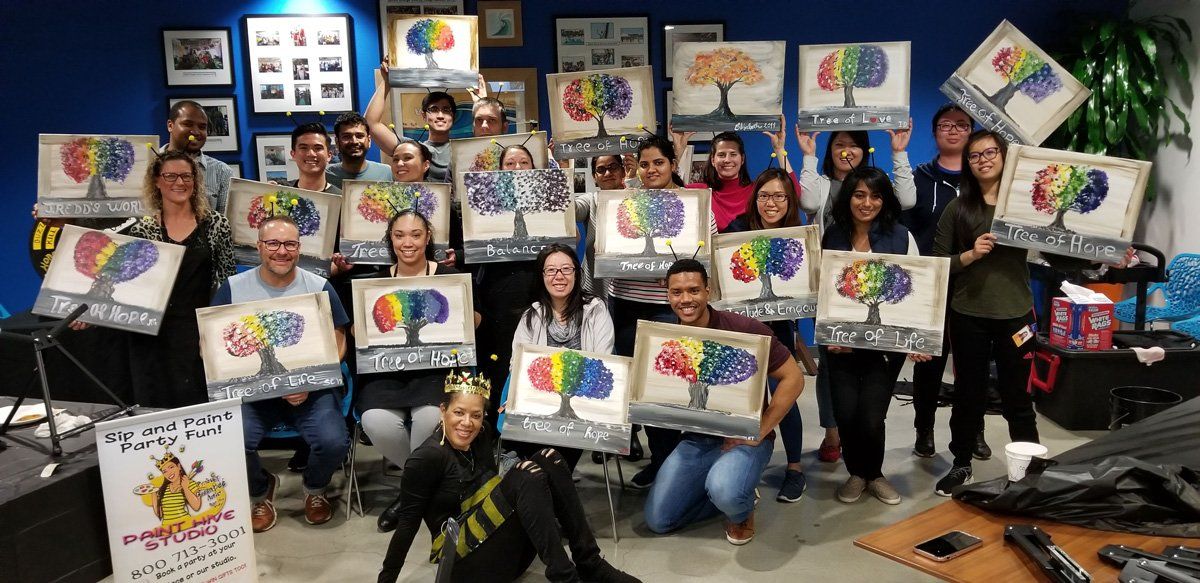Group of people holding up paintings of colorful trees, smiling in a room with blue walls and paintings.