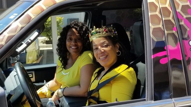 Two women smiling in a vehicle decorated with giraffe print. One wears a crown.