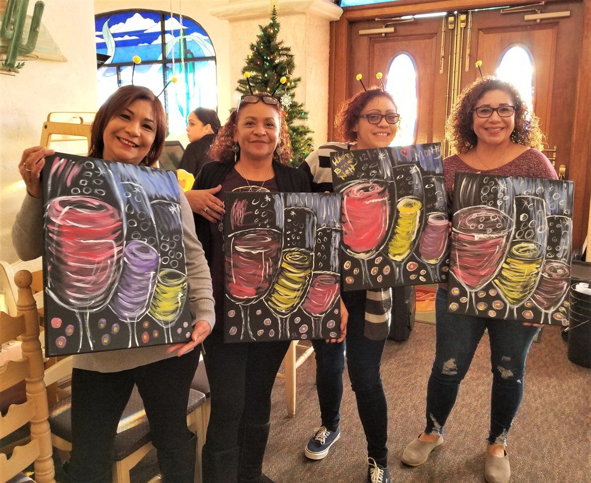 Four smiling people hold up paintings of wine glasses in a restaurant setting.