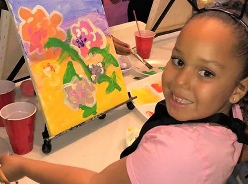 Young girl smiles while painting flowers on canvas.