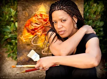 Woman holding paintbrushes sits in front of her abstract art.