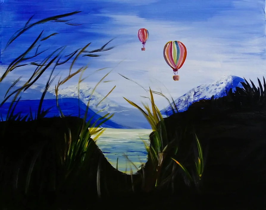Two hot air balloons over a lake and mountains, viewed from grassy foreground.