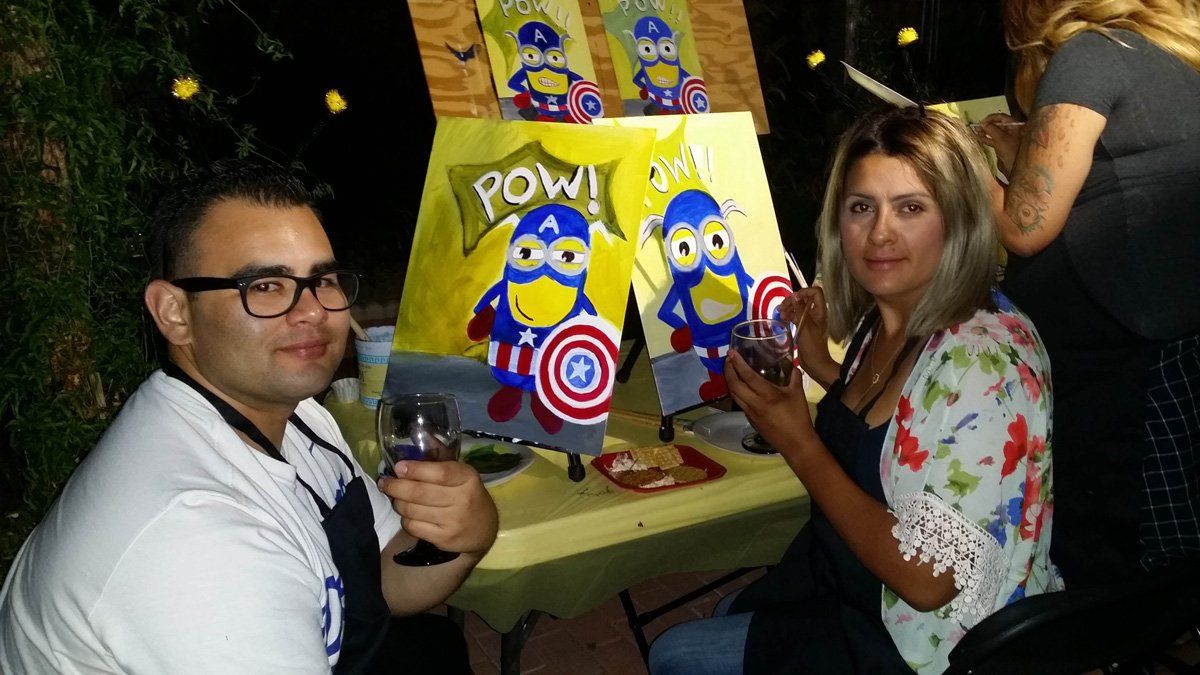 Two people paint Captain America/Minion art at an outdoor party. They hold drinks, and a sign says 