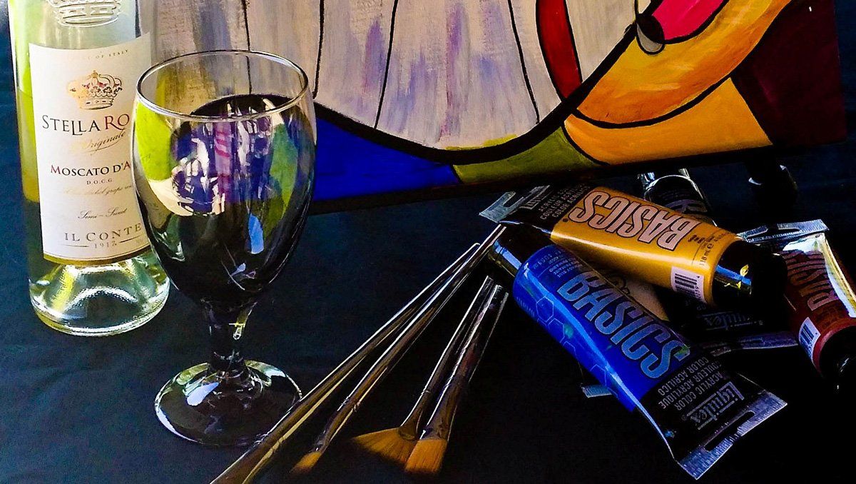 Wine glass and bottle with paint tubes, brushes, and abstract colorful painting on canvas.