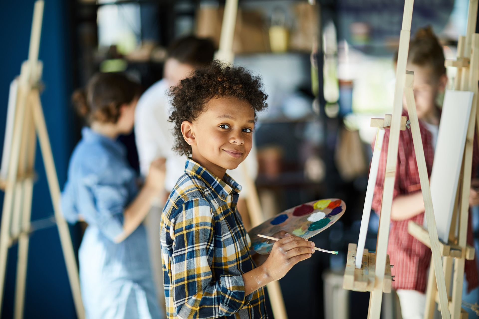 Boy holding a paintbrush and palette, smiling, in an art class setting, surrounded by easels.