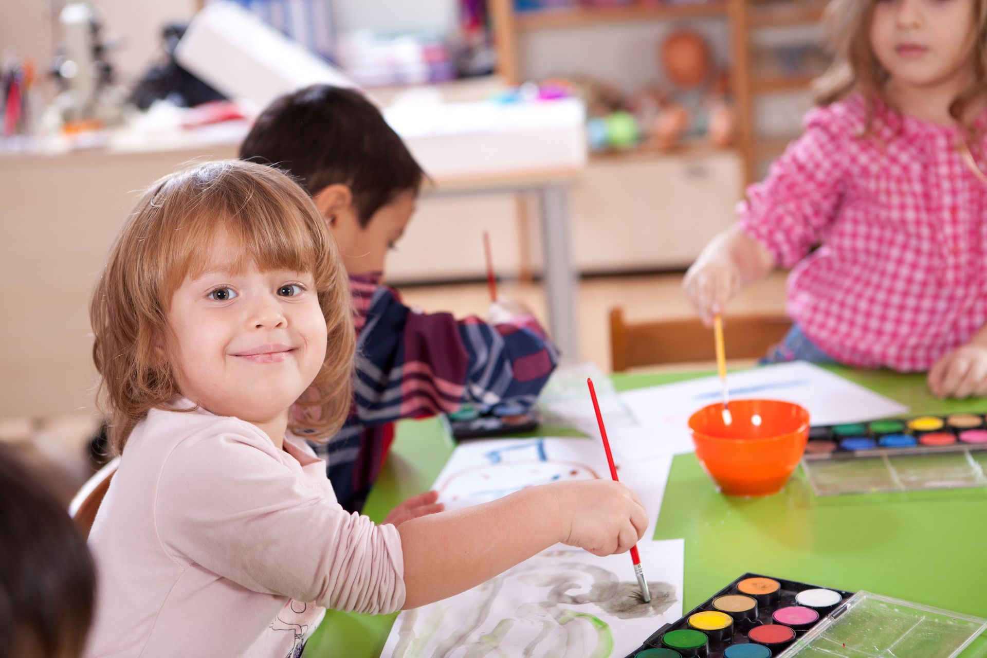 Children painting at a table, smiling. Watercolor paints, brushes, and paper. Classroom setting.