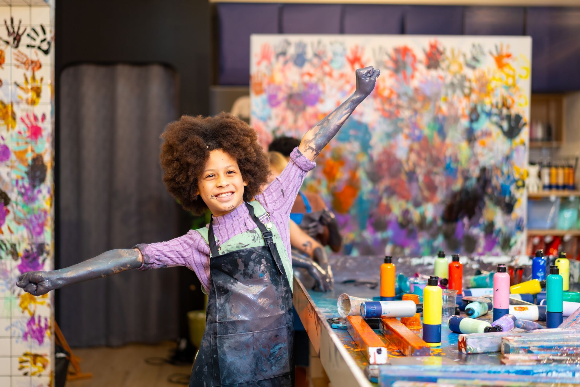 Child in art studio with arms covered in paint, smiling with hands up in front of a colorful handprint backdrop.
