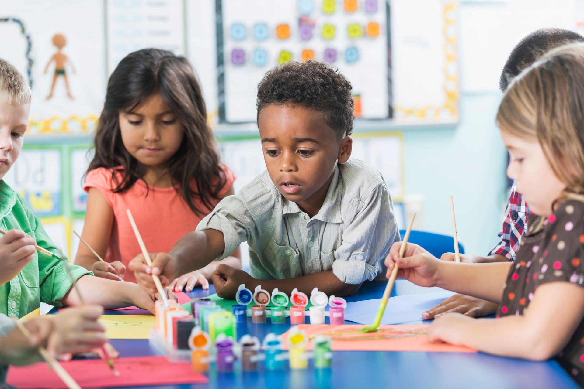 Children painting at a table in a classroom with paintbrushes and colorful paints.