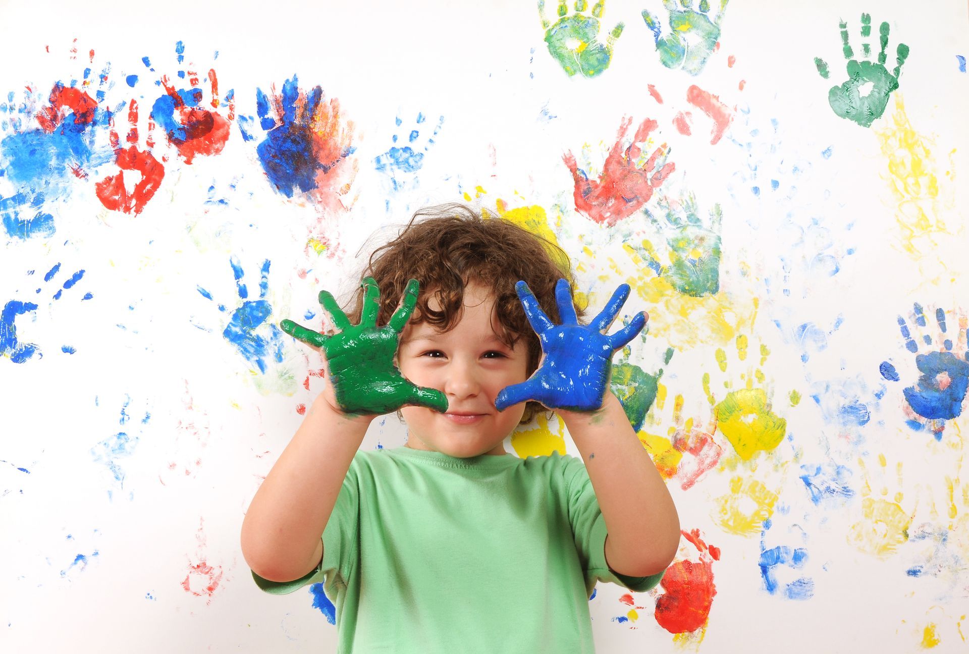 Boy with green and blue paint on his hands smiles in front of a wall covered in colorful handprints.