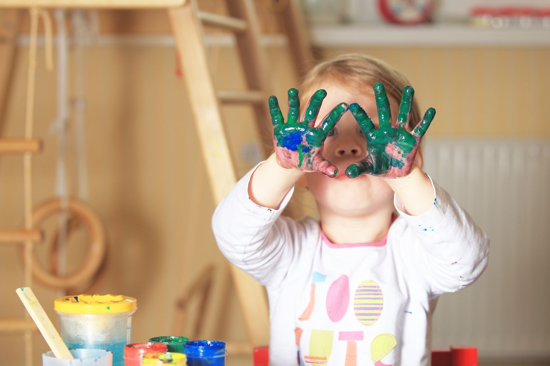 Child with paint-covered hands looking through them. Paints and play structure in background.