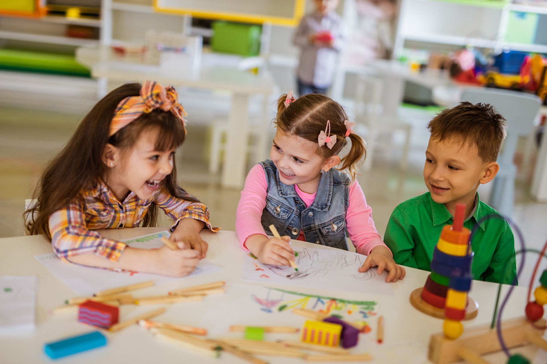 Three children at a table, smiling and coloring. Classroom setting with crayons and toys visible.