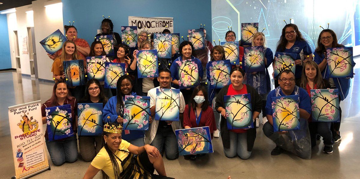 Group of people holding up paintings in front of a blue wall. Paintings feature yellow and white designs.
