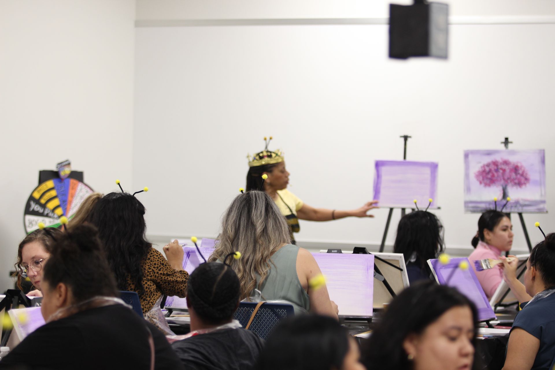People paint at easels; instructor in a bee-themed costume points to a purple painting, room with white walls.