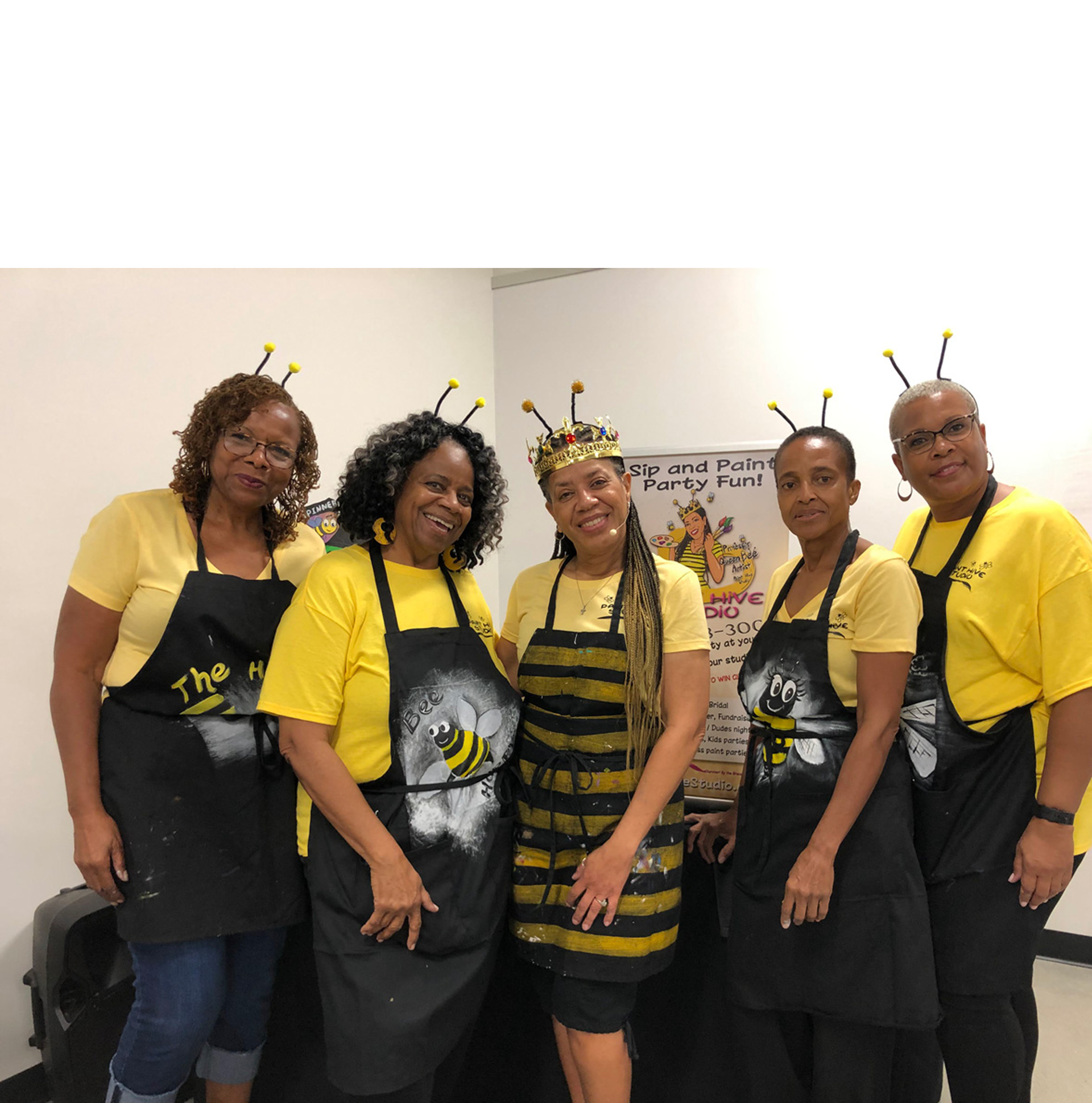 Five people in yellow shirts, black aprons with bee designs, and bee antennae stand near a sign.