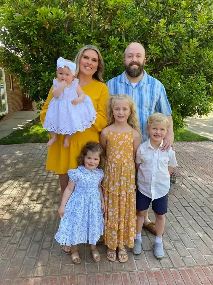 Family of five posing outdoors on a brick path, with three young children in front and a baby being held.