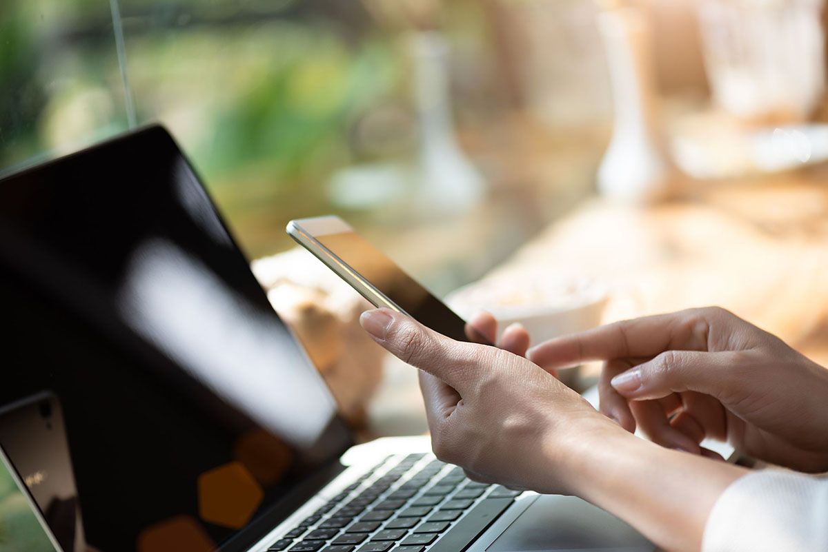 Hands typing on a smartphone beside a laptop in a blurred office setting