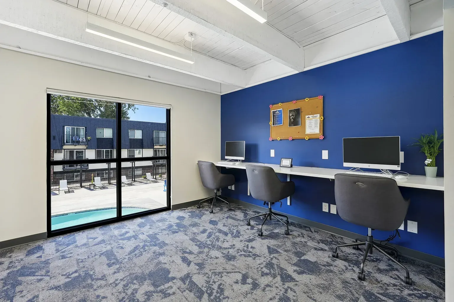 A modern study room with blue walls, three chairs, desk space, computers, a corkboard, and a large window facing a pool at Avec Greeley.