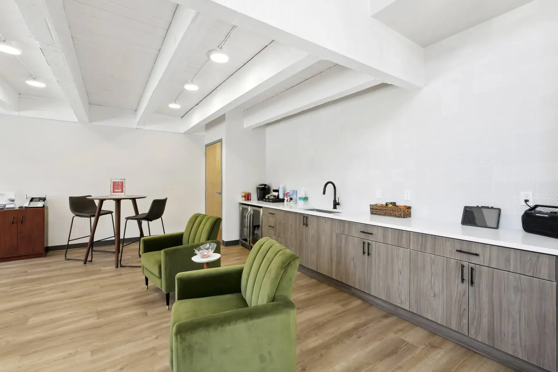 A modern breakroom with two green velvet chairs, a wood-toned counter with a sink, and a small bistro table in the corner at Avec Greeley.