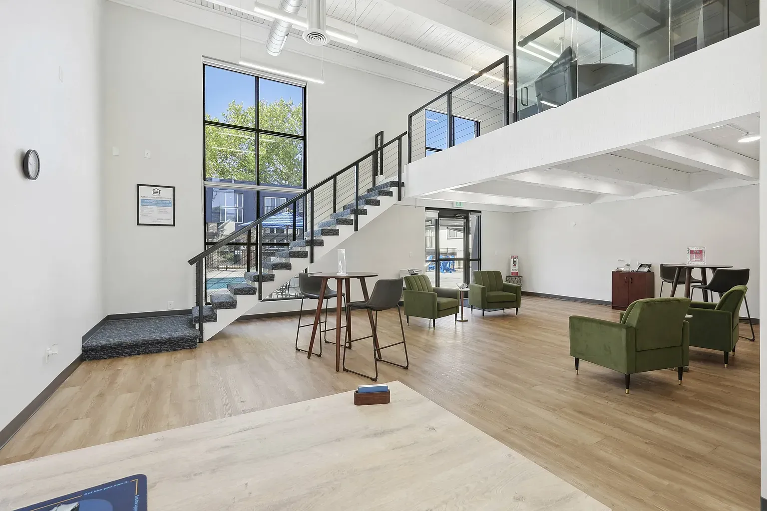Modern, open-plan office lobby with light wood floors, a metal staircase, tall windows, and several olive green chairs.