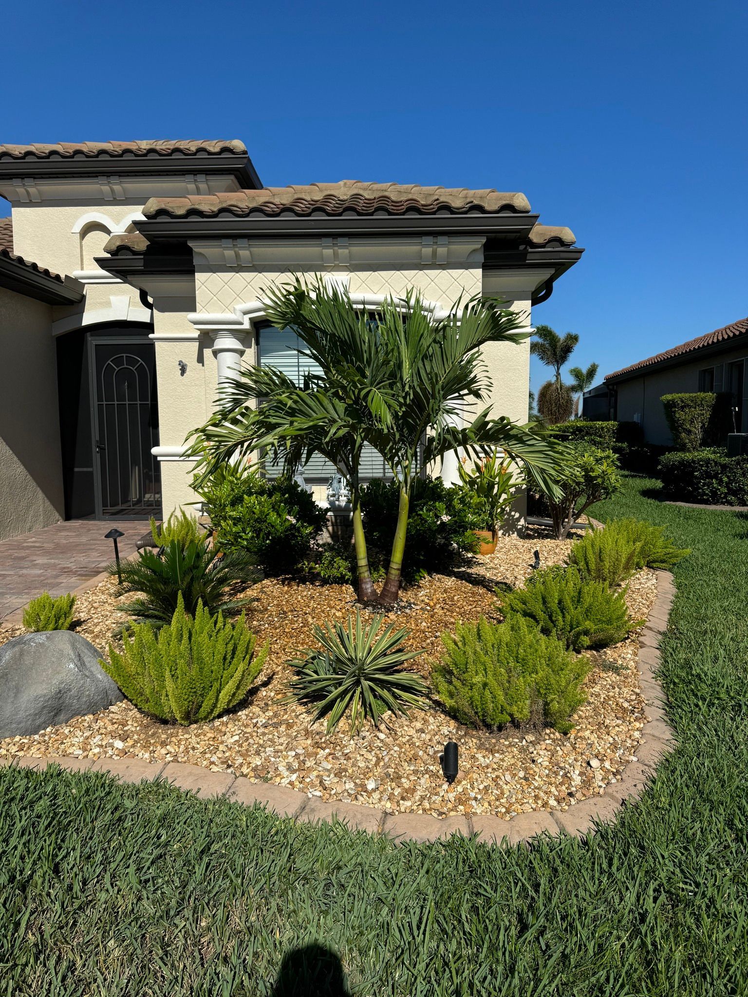 Front yard with tropical plants and gravel landscaping beside a stucco house under a clear blue sky