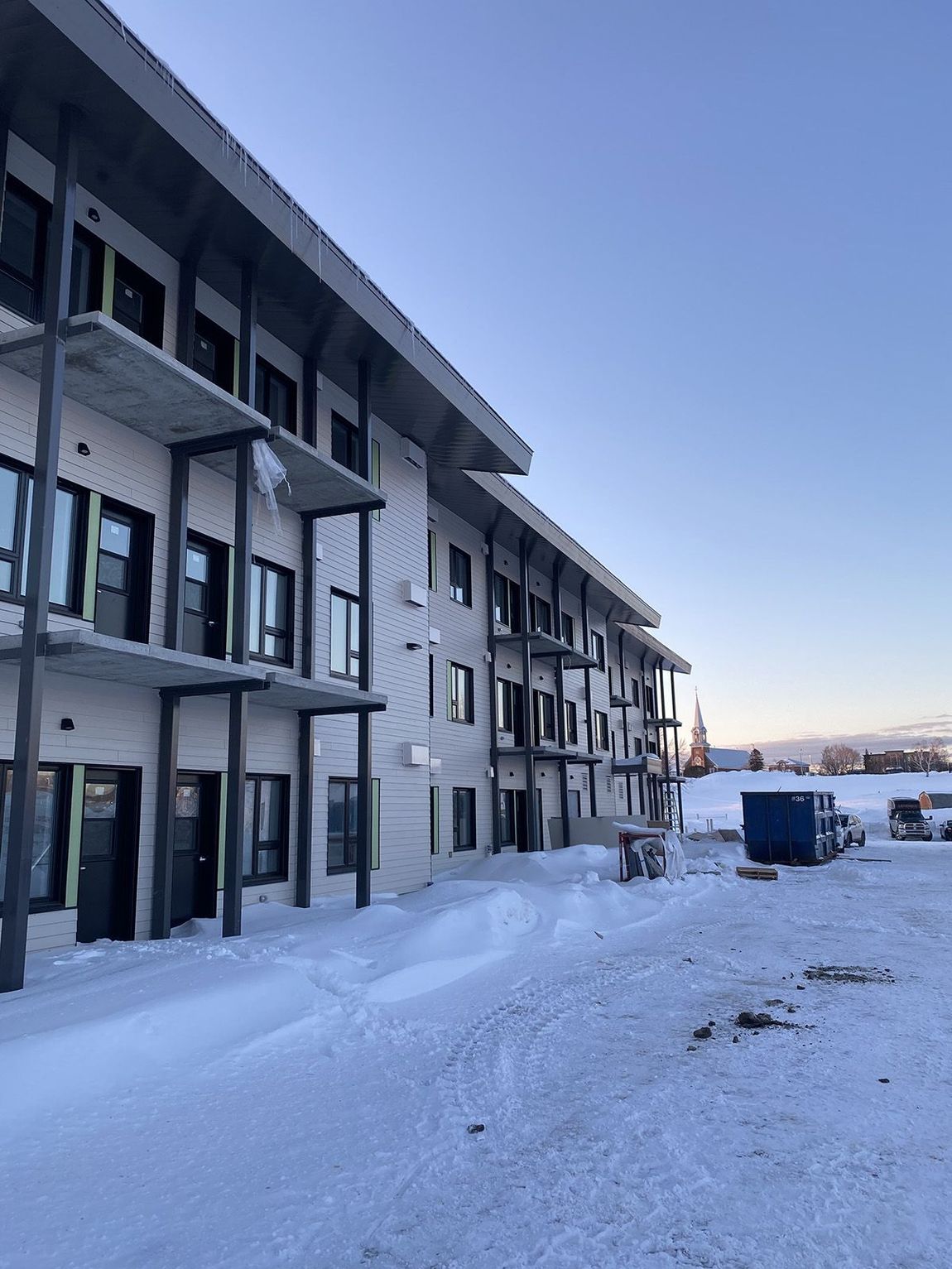 Immeuble de trois étages avec balcons, façade en briques, dans un paysage enneigé sous un ciel dégagé.