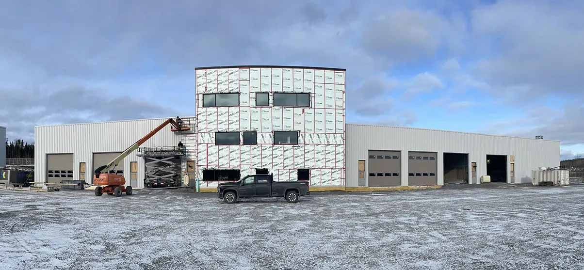 Un bâtiment en construction, à la façade blanche, avec un camion noir garé devant. Sol enneigé, ciel bleu.