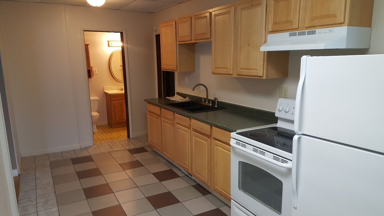 Kitchen with light wood cabinets, white appliances, and a doorway to a bathroom.