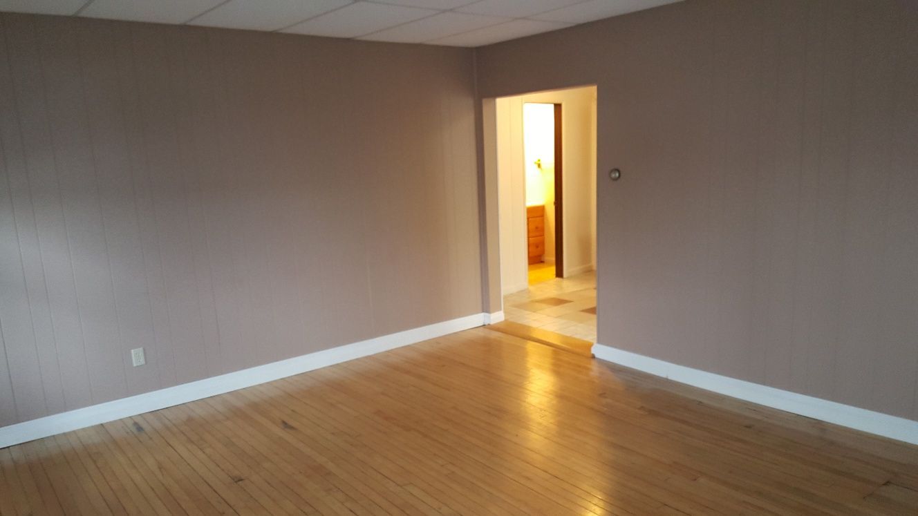 Empty room with wooden floor, tan walls, and a doorway leading to a lit room.