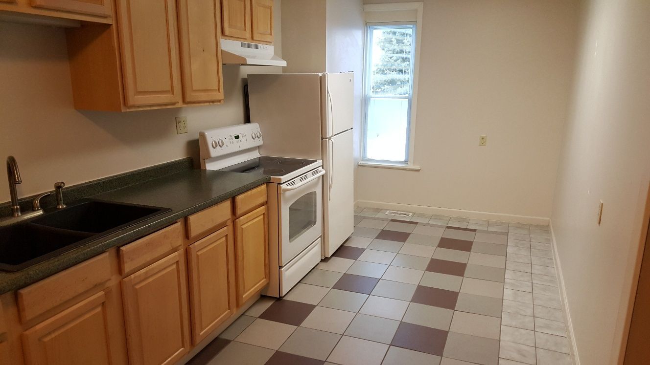 Kitchen with wood cabinets, dark countertop, white appliances, and checkered floor.