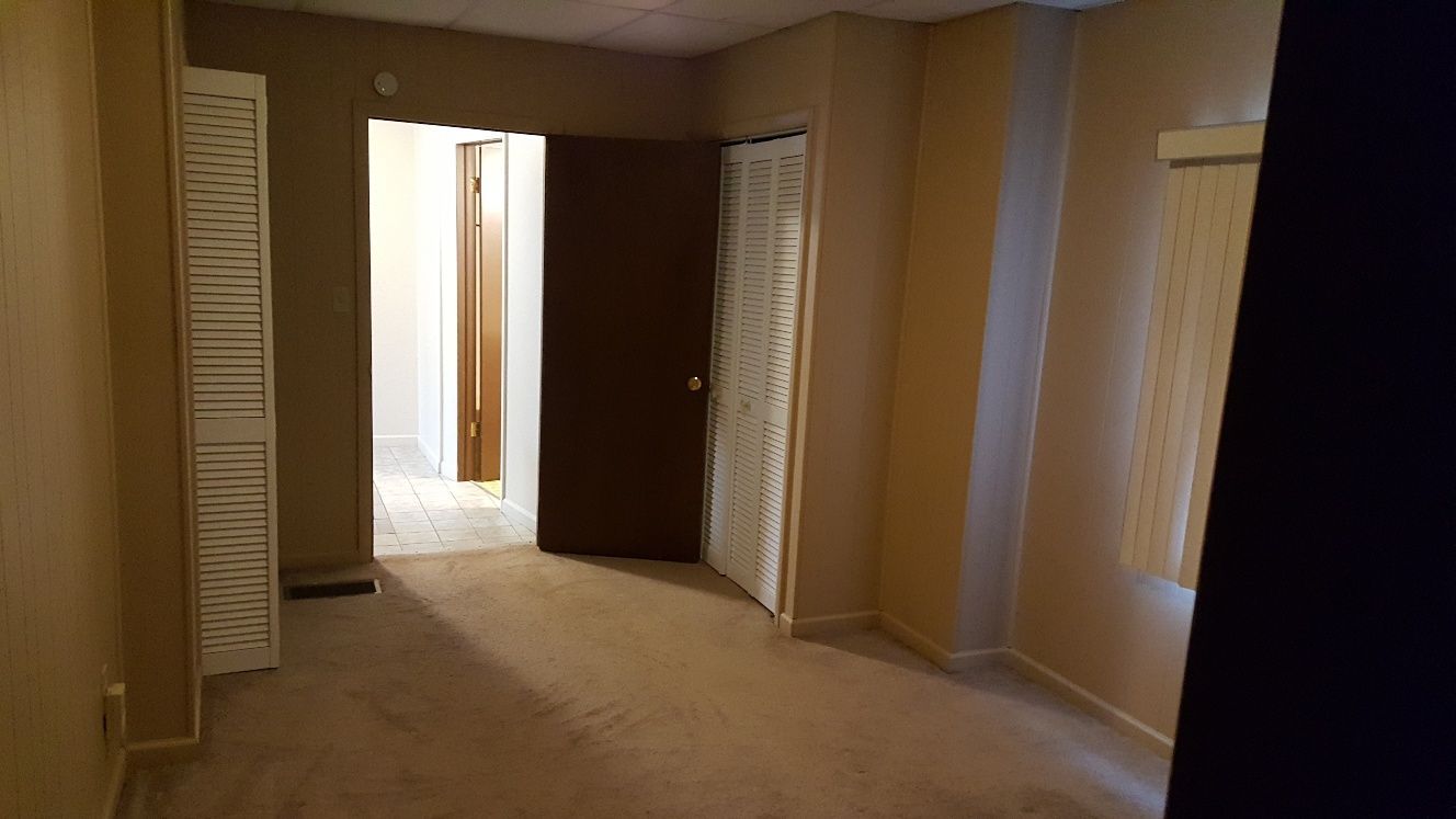 Empty bedroom with beige walls, carpet, and a partially open brown door leading to a hallway.