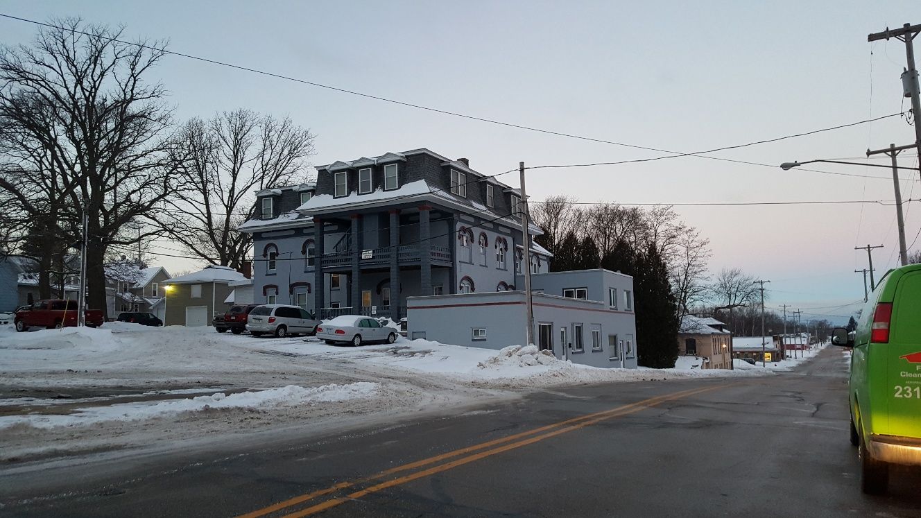 Snowy street scene with a three-story building, bare trees, and a green van.