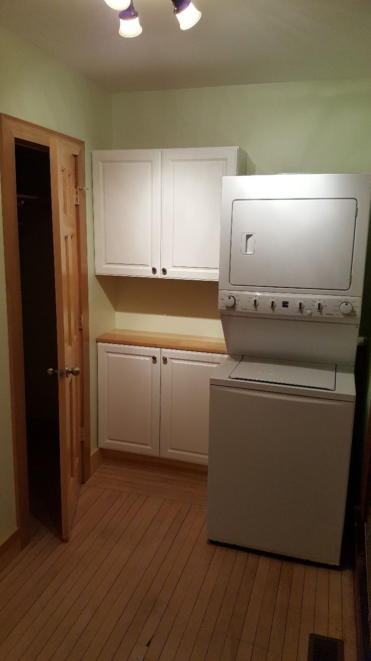 Laundry room with stacked white washer/dryer, white cabinets, wooden countertop and floors.