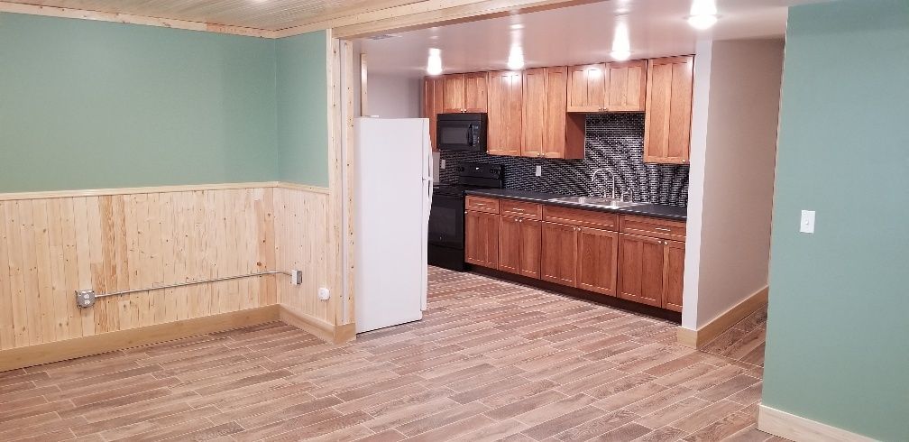 Interior view of a kitchen and living area, featuring light wood cabinets, black appliances, and pale green walls.