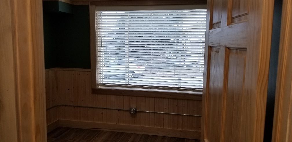 Interior view of a small room with a window and wooden door, walls and floor. Window blinds are closed.