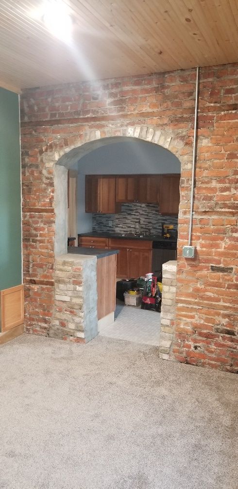 An arched brick doorway leads to a kitchen. Brick walls and a wood ceiling are visible.