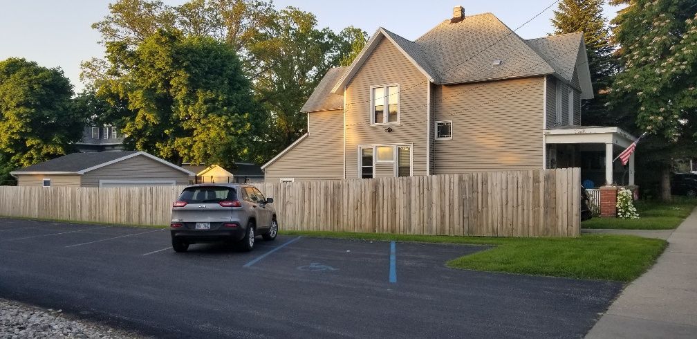 A car parked in front of a house behind a wooden fence. The house has a porch and American flag.