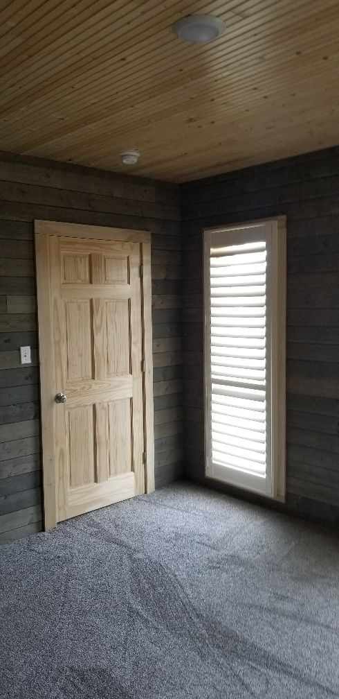 Room with wood-paneled walls and ceiling, a door, a window with shutters, and gray carpet.