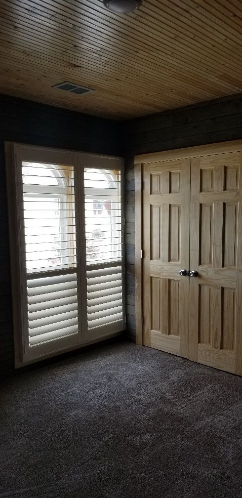 Room interior with window shutters, wooden doors, and wood-paneled ceiling, dark carpet.