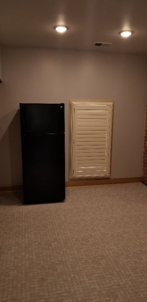 Black refrigerator next to a small, shuttered window in a room with tan carpet and a neutral wall.