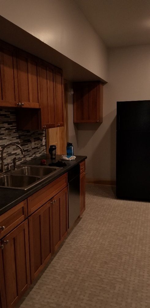 Kitchen with brown cabinets, black countertops, and stainless steel sink and appliances. Black refrigerator. Carpet flooring.