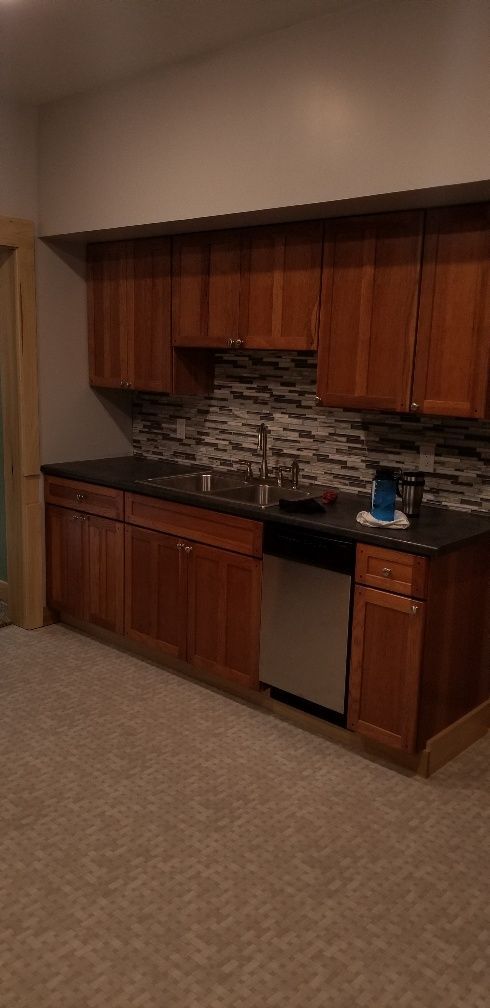 Kitchen cabinets, sink, and dishwasher against a mosaic tile backsplash, black countertop, carpet flooring.